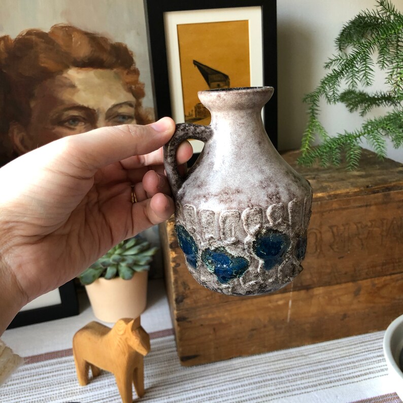May include: A small, white ceramic vase with a blue and green glaze. The vase has a rounded body and a handle. It is decorated with a repeating pattern of stylized flowers.