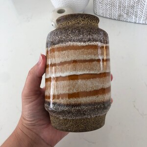 May include: A cylindrical ceramic vase with horizontal bands of brown, tan, and white. The vase has a textured, speckled base and a slightly flared rim. The color palette is earthy and neutral, suitable for various decor styles.