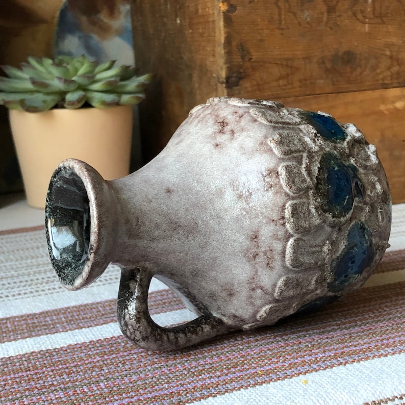 May include: A white ceramic vase with a textured surface and blue and brown accents. The vase has a rounded body and a handle.