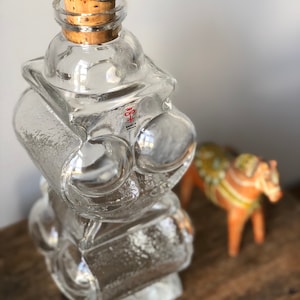 May include: A clear glass bottle with a textured surface and a cork stopper. The bottle has a silver ring on the top and a red logo that says "Kosta Boda" on the side.