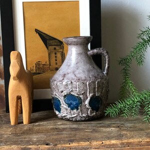 May include: A ceramic vase with a white glaze and blue accents. The vase has a textured surface and a rounded body with a wide mouth and a handle.