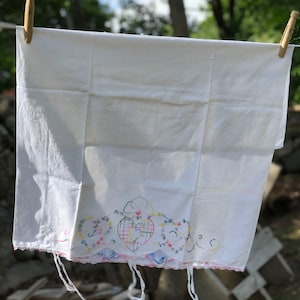 Puede incluir: Toalla de té de algodón blanco con un diseño de corazón bordado de colores y un borde festoneado. La toalla está colgada de un tendedero con pinzas de madera.