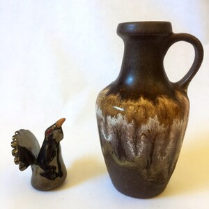 May include: A brown ceramic vase with a white and brown glaze pattern. The vase has a rounded body and a narrow neck with a handle. Next to the vase is a small ceramic figurine of a bird with a brown and black body and a gold tail.