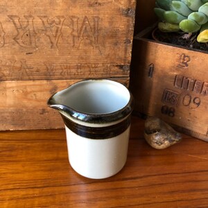 May include: A small, white ceramic creamer with a brown and black band around the top. The creamer has a rounded shape and a small spout.