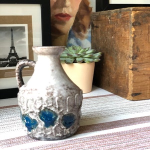 May include: A white ceramic pitcher with blue accents. The pitcher has a textured surface and a handle. It is sitting on a striped tablecloth.