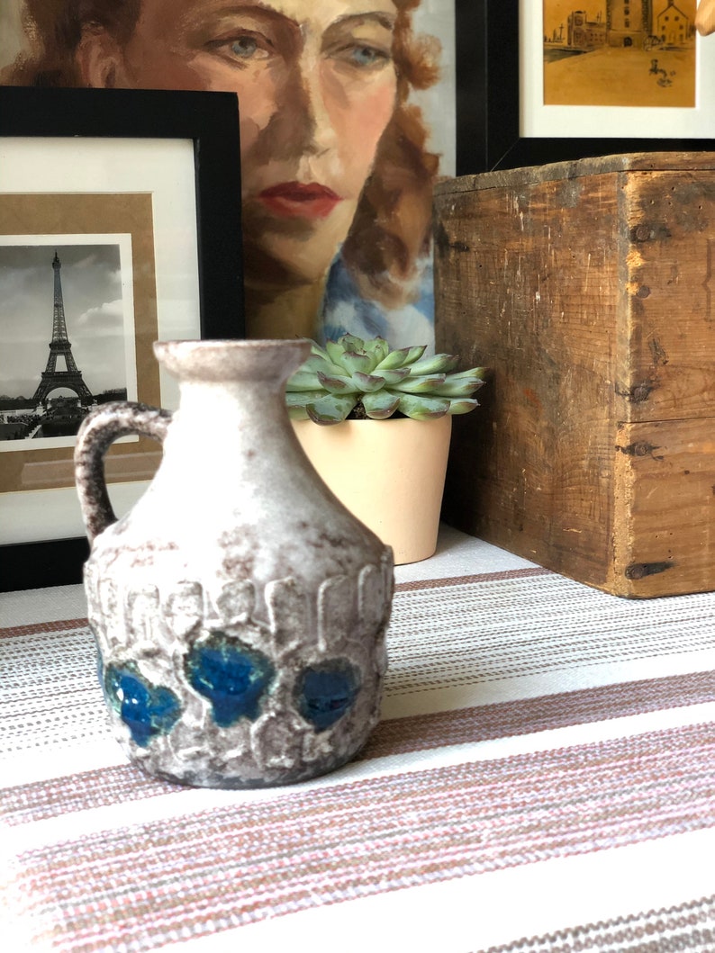 May include: A white ceramic pitcher with blue accents. The pitcher has a textured surface and a handle. It is sitting on a striped tablecloth.