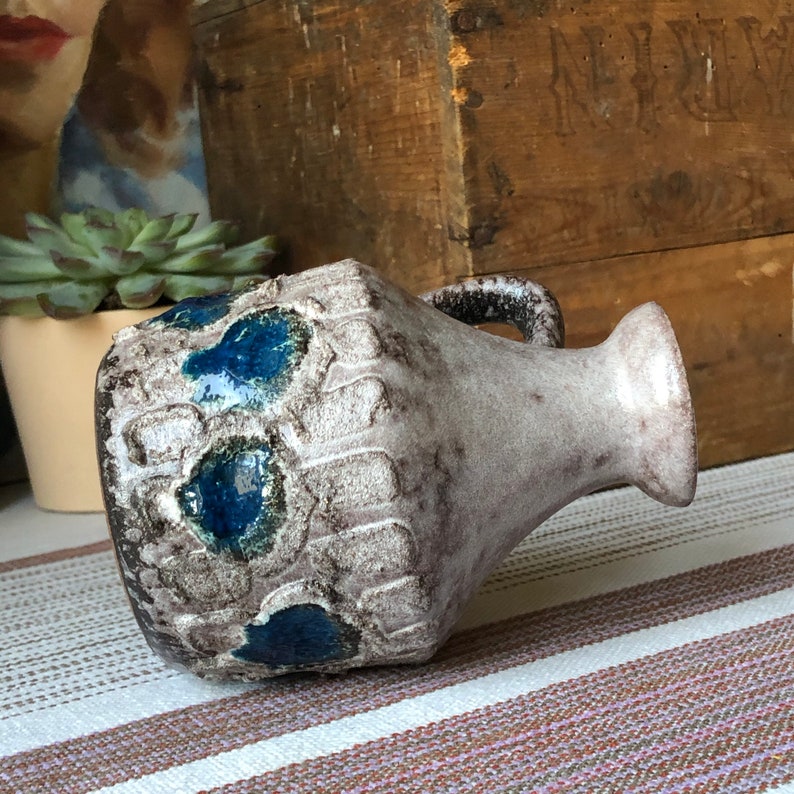 May include: A white ceramic vase with blue and brown glaze. The vase has a textured surface and a handle. It is a vintage style vase.
