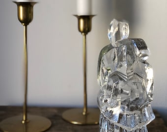 May include: A clear glass sculpture with a geometric design sits on a wooden surface in front of two gold candle holders with white candles.