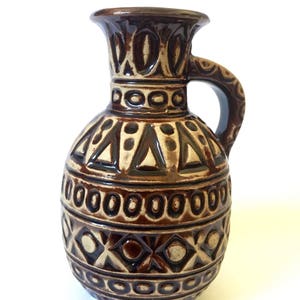 May include: A brown ceramic pitcher with a handle and a wide mouth. The pitcher features a repeating geometric pattern in a lighter brown color. The design includes triangles, circles, and diamond shapes. The pitcher is likely used for decorative purposes.
