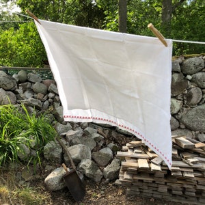 Puede incluir: Un mantel de lino blanco con un borde bordado rojo cuelga de un tendedero al aire libre. El mantel está drapeado sobre una pila de madera y piedras.