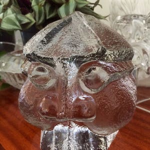 Puede incluir: Una escultura de vidrio transparente de una cara con una superficie texturizada. La cara tiene una nariz grande, pómulos prominentes y una boca ancha. La escultura está sentada sobre una superficie de madera.