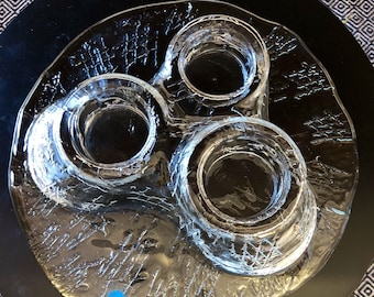 May include: A clear glass candle holder with three circular openings. The holder has a textured surface and is sitting on a black surface.
