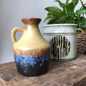 May include: A brown and yellow ceramic vase with a blue and black glaze. The vase has a wide mouth and a handle. It is sitting on a wooden surface next to a white ceramic planter with a brown design.
