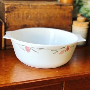 Op de afbeelding: Witte keramische mengkom met een bloemenmotief met roze bloemen en grijze stelen. De kom heeft een afgeronde vorm met twee handvatten aan de zijkanten.