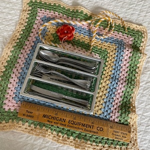 May include: A vintage flatware set in a clear plastic tray, resting on a colorful, crocheted placemat. The set includes spoons, forks, and knives. A wooden ruler with text from Michigan Equipment Co. is also present.