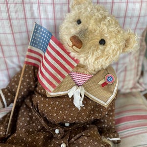 May include: A vintage teddy bear wearing a brown dress with white star patterns and a patriotic collar. The bear holds a small American flag. The bear's fur is a light beige color, and the dress has white buttons.