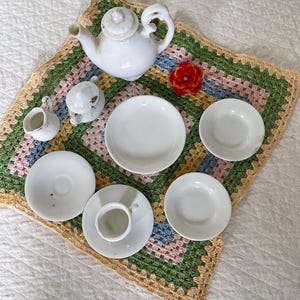 May include: A white porcelain tea set, including a teapot, creamer, sugar bowl, cup and saucer, and several plates, arranged on a colorful crocheted mat. A red flower adds a pop of color to the scene. The set is ideal for a dollhouse or display.
