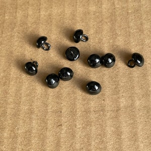 May include: A collection of ten black buttons with a shiny finish. The buttons are round and have a small loop on the back for sewing.