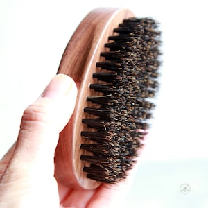 May include: A wooden oval-shaped brush with black bristles. The brush is held in a hand.