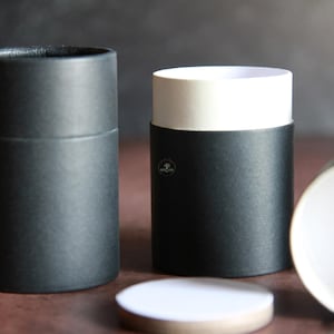 May include: Several cylindrical black paper tubes with white interiors and lids are displayed. One tube is open, revealing the white interior. The tubes are arranged on a brown surface, suggesting a packaging or storage theme.