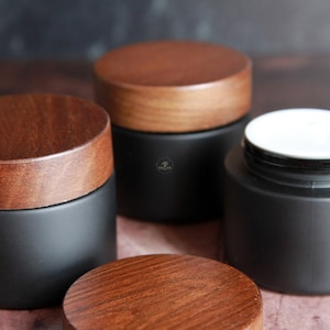 Puede incluir: Varios tarros cosméticos negros con tapas de madera. Un tarro está abierto, revelando un interior blanco. Los tarros son cilíndricos y tienen un acabado mate. Las tapas de madera son redondas y tienen vetas naturales.
