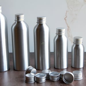 Puede incluir: Una colección de botellas de aluminio plateado de varios tamaños, con tapas de rosca a juego. Las botellas están dispuestas en fila, con varias tapas delante. Las botellas tienen un acabado metálico liso.