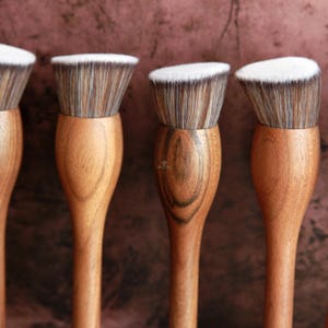 May include: Five makeup brushes with light brown wooden handles and white and brown bristles. The brushes have a rounded shape at the base and a slightly angled brush head. The bristles are white-tipped.