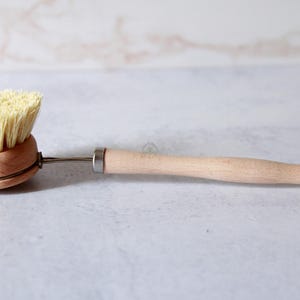 May include: A dish brush with a round wooden head, light-colored stiff bristles, and a long, smooth wooden handle. A thin metal rod connects the brush head to the handle, and a small metal loop is attached to the end of the handle for hanging.