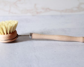 May include: A dish brush with a round wooden head featuring light-colored stiff bristles, a long wooden handle, and a metal connector. A small metal loop is attached to the end of the handle for hanging the cleaning tool.