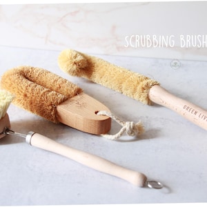 May include: A scrubbing brush set featuring three cleaning tools with light wood handles and tan bristles. One brush has a round head, another is oval-shaped, and the third is long and cylindrical. Each handle includes a hanging loop and text.