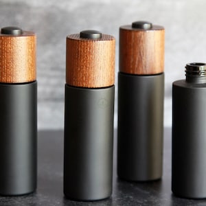 May include: Four black cylindrical bottles with wooden tops. Three bottles have a black cap on top of the wooden top. One bottle is open. The bottles are arranged on a dark surface.