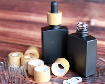May include: Two black glass bottles with bamboo lids and droppers, along with several extra bamboo lids and glass droppers. The bottles and lids are on a wooden surface.