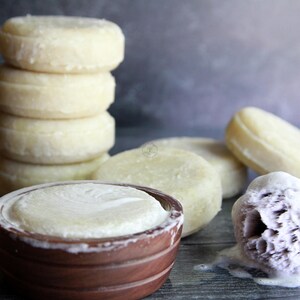 Puede incluir: Una pila de jabones redondos y claros junto a un cuenco de madera con jabón y una brocha de afeitar con espuma. Varios otros jabones están esparcidos alrededor del cuenco. Los artículos están dispuestos sobre una superficie oscura.