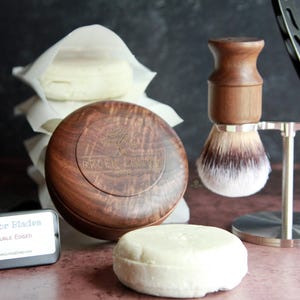 May include: A collection of grooming products, including a wooden shaving brush, a straight razor, and a wooden soap dish. The image also shows a stack of wrapped soaps and a tin of razor blades.