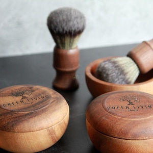 May include: A collection of wooden shaving accessories, including a shaving brush, a bowl, and two containers with lids. The containers and bowl have the words "GREEN LIVING" and a tree logo. The brush has a dark bristle head.
