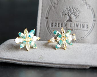 May include: Two gold-toned rings featuring a flower-shaped design with marquise-cut light blue and clear crystals. The rings are displayed in front of a gray fabric pouch embossed with a tree logo and the text GREEN LIVING.