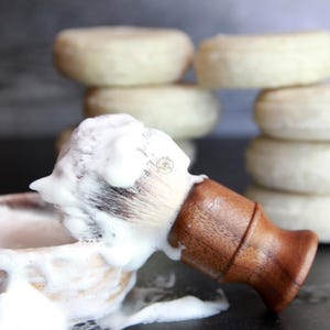 Puede incluir: Una brocha de afeitar con mango de madera está cubierta de crema de afeitar blanca, junto a un cuenco de afeitar con más crema. Varias pastillas de jabón redondas y blanquecinas están apiladas en el fondo. La escena sugiere un producto de afeitado.