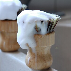 May include: Two wooden shaving brushes with white shaving cream on the bristles. The brushes are in a bathroom sink.