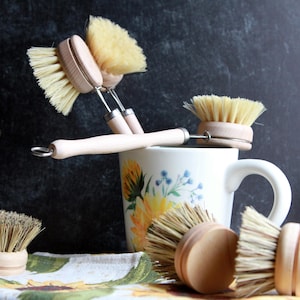 May include: A set of five wooden-handled dish brushes with natural bristles. The brushes are arranged in a group, with some leaning against a white mug with a floral design. The mug is sitting on a white and brown patterned cloth.