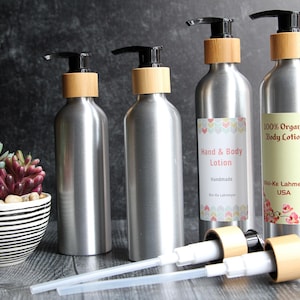 Puede incluir: Botellas de loción plateadas con bombas negras y cuellos de bambú. Dos botellas tienen etiquetas que dicen "Hand & Body Lotion" y "100% Organic Body Lotion". Una planta suculenta decorativa en una maceta a rayas está en primer plano.