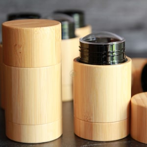 Può includere: Diversi contenitori cilindrici in bambù color chiaro sono esposti. Alcuni hanno coperchi, mentre altri sono aperti, rivelando un interno nero. Un contenitore ha un coperchio trasparente a cupola. I contenitori sono disposti su una superficie scura.