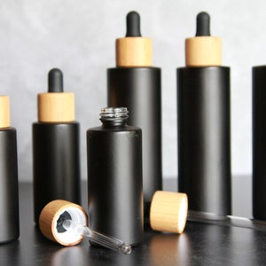 May include: Six black glass bottles with bamboo lids and droppers. The bottles are all the same size and shape, and they are arranged in a row on a gray surface. One of the bottles is open and the dropper is lying on the surface.