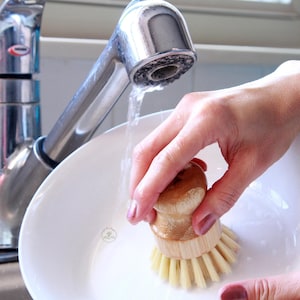 May include: A person rinsing a wooden dish brush with a natural bristle brush head under running water in a white ceramic sink.