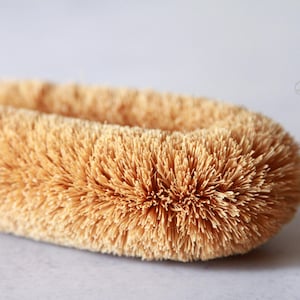 May include: Close-up of a natural bristle brush with a wooden handle. The bristles are made of coconut fibers and are a light brown color.