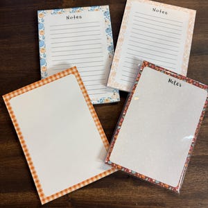 May include: Four rectangular notepads with various borders. Two pads feature lined paper with the word Notes printed at the top, one with blue pumpkins and one with orange floral patterns. Two pads have blank dotted paper, one with an orange gingham border and one with a dark autumn leaf border.