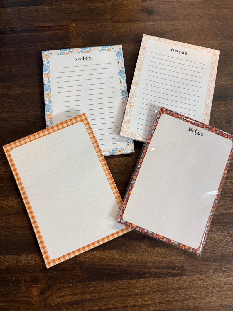 May include: Four rectangular notepads with various borders. Two pads feature lined paper with the word Notes printed at the top, one with blue pumpkins and one with orange floral patterns. Two pads have blank dotted paper, one with an orange gingham border and one with a dark autumn leaf border.