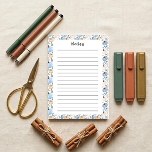 May include: A rectangular notepad with the word Notes printed at the top, featuring horizontal lines and a border of blue and orange pumpkins. The notepad is surrounded by three pens, three markers, gold scissors, and three bundles of cinnamon sticks.