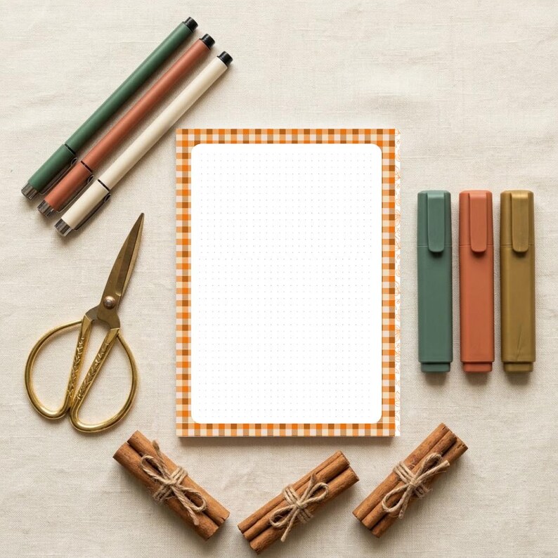 May include: A rectangular notepad with a white dot grid center and an orange and white gingham border, accompanied by three pens, three markers, gold-toned scissors, and three bundles of cinnamon sticks tied with twine.