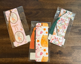 May include: Three rectangular paper bookmarks in clear plastic sleeves. The left bookmark features ducks in oval frames with a gold tassel. The center bookmark shows orange cats, pink ghosts, and stars with an orange tassel. The right bookmark displays mushrooms and autumn leaves with a dark green tassel.