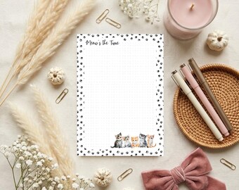 May include: A rectangular notepad with a white dotted grid background, a border of black paw prints, and five kittens at the bottom. The text Meow's the Time is printed in black at the top left corner of the page, which is centered on a surface.
